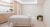 A tranquil treatment room featuring a massage table dressed in neutral linens with white bolster pillows. The space includes light wood cabinetry, a sleek sink area, and decorative potted plants, embodying a modern, clean aesthetic perfect for therapeutic services.