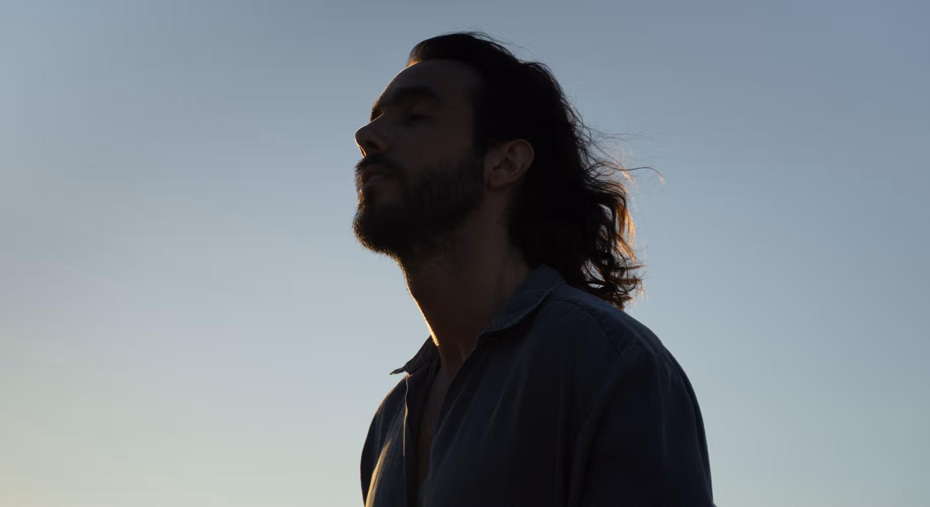 Man with long hair and beard looking up, covered by a shadow, against a blue sky