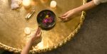 Two light skin arms and hands extend over a golden table with candles, an incense in an incense holder and a large, white crystal. They are each holding a burnt Santo Palo large stick over a bowl of water with flowers floating.