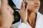 A woman dressed in a white robe applies lotion to her face.