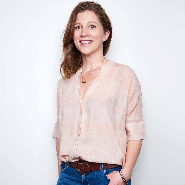 A white woman in a pink blouse, brown belt, dark blue denim and a gold necklace. They have brown hair, blue eyes and pink lipstick. They have one arm behind their back and another hand in their jean pocket, smiling with teeth looking forward.