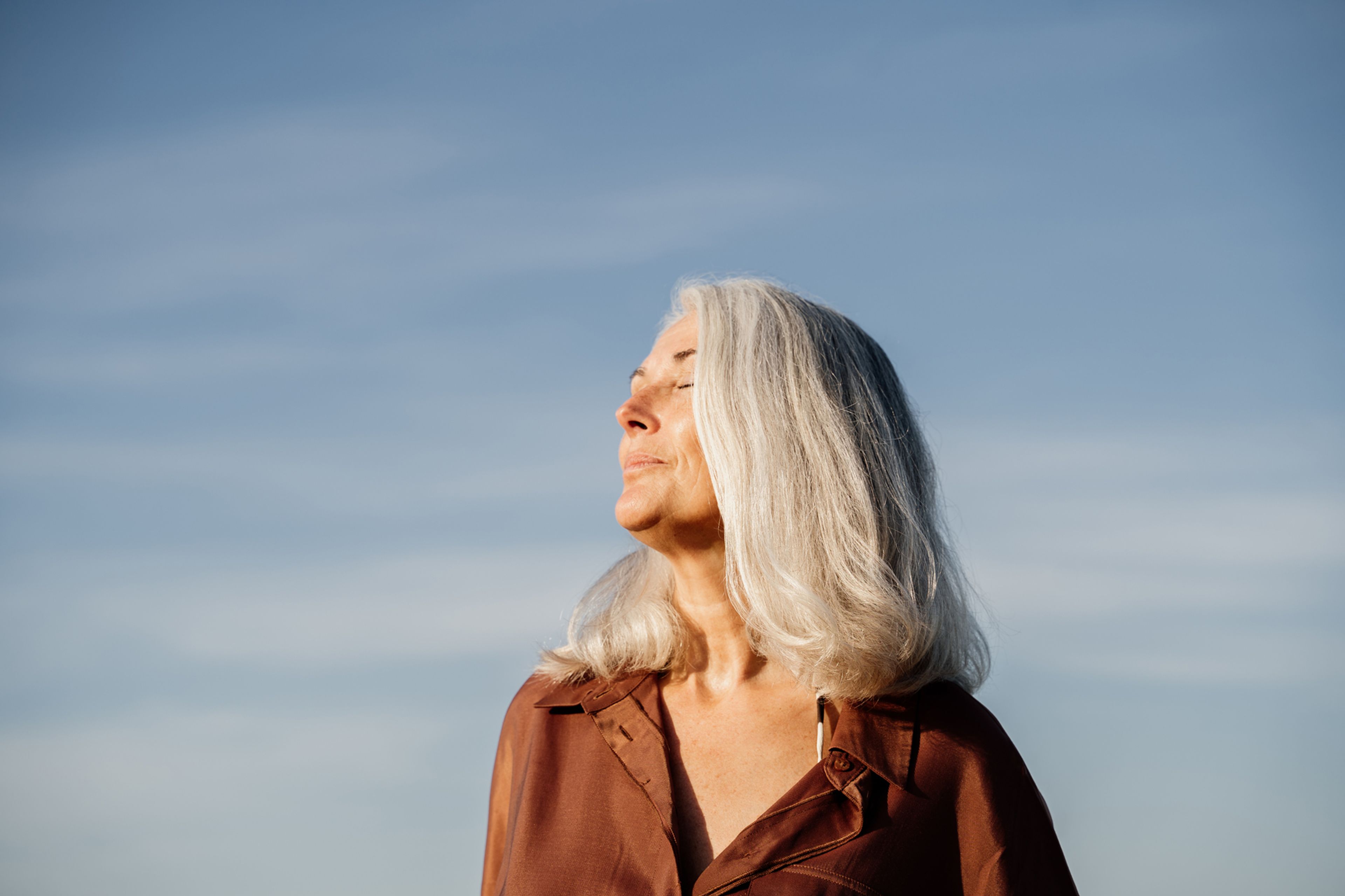 Grey haired woman with eyes closed, standing in front of a blue sky