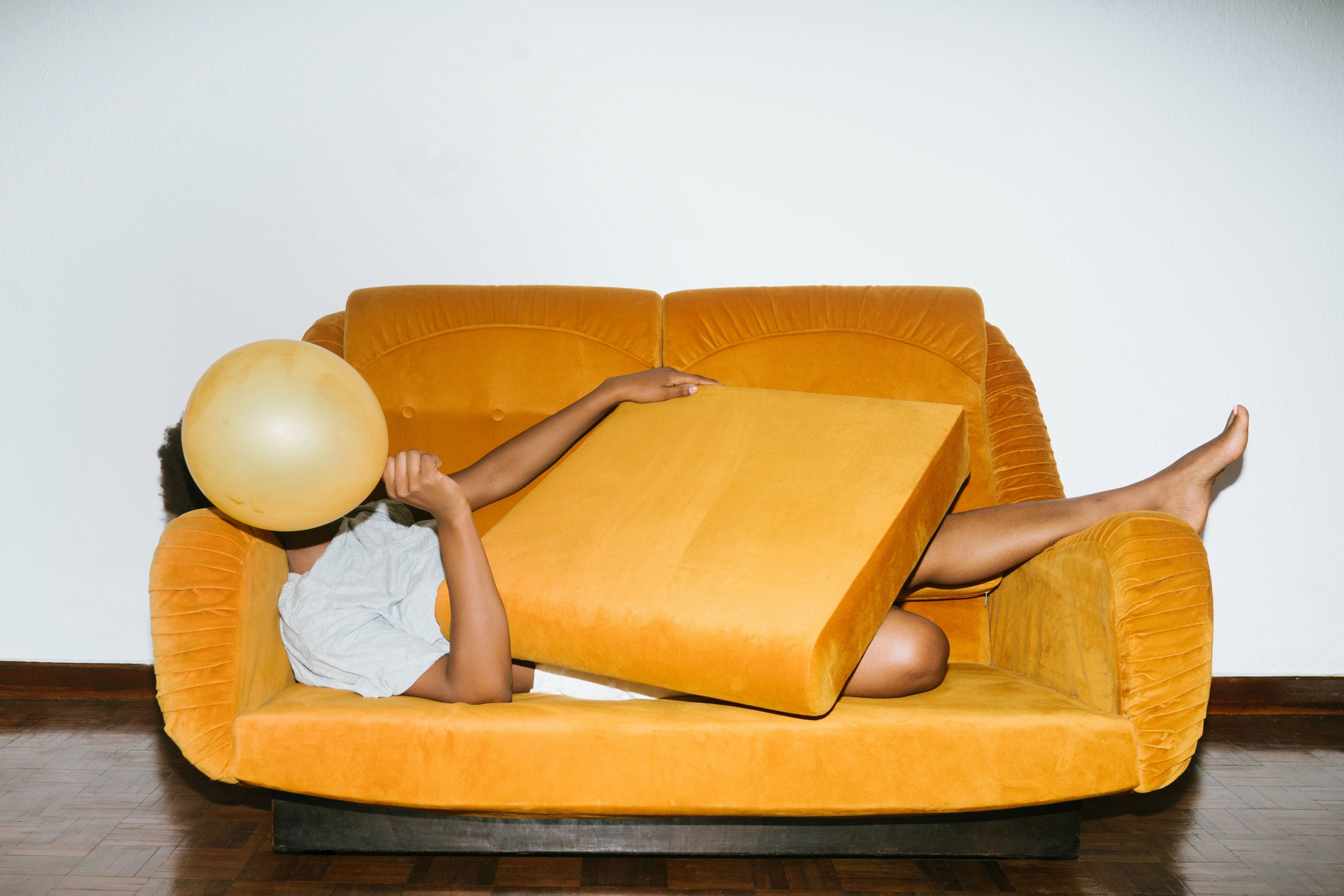A brown woman laying on an orange sofa covers her face with an orange balloon.