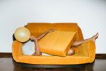 A brown woman laying on an orange sofa covers her face with an orange balloon.