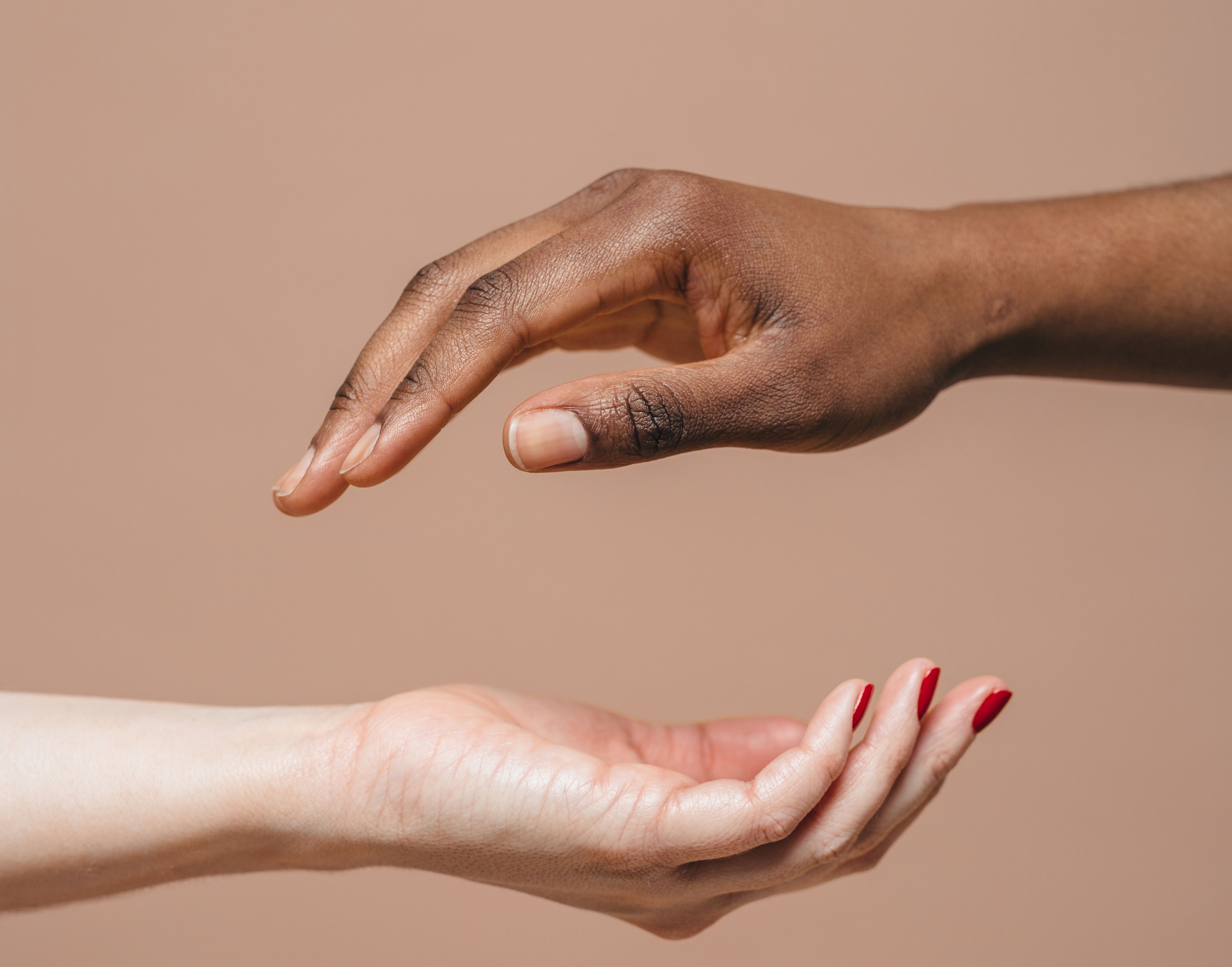 Two hands hover over each other, one above the other. The bottom hand is white with red nail polish and the hand above is brown with bare nails.  