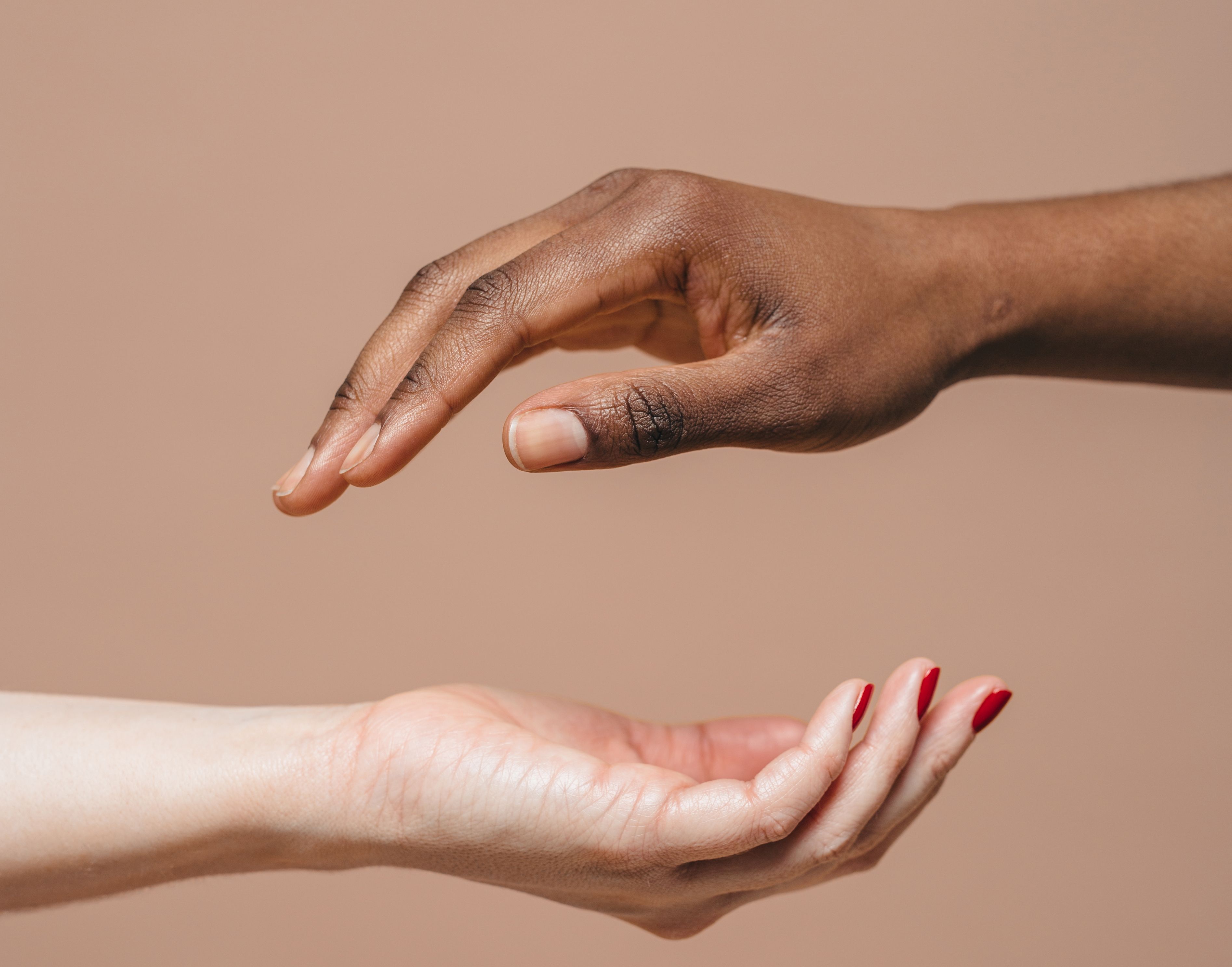 Two hands hover over each other, one above the other. The bottom hand is white with red nail polish and the hand above is brown with bare nails.