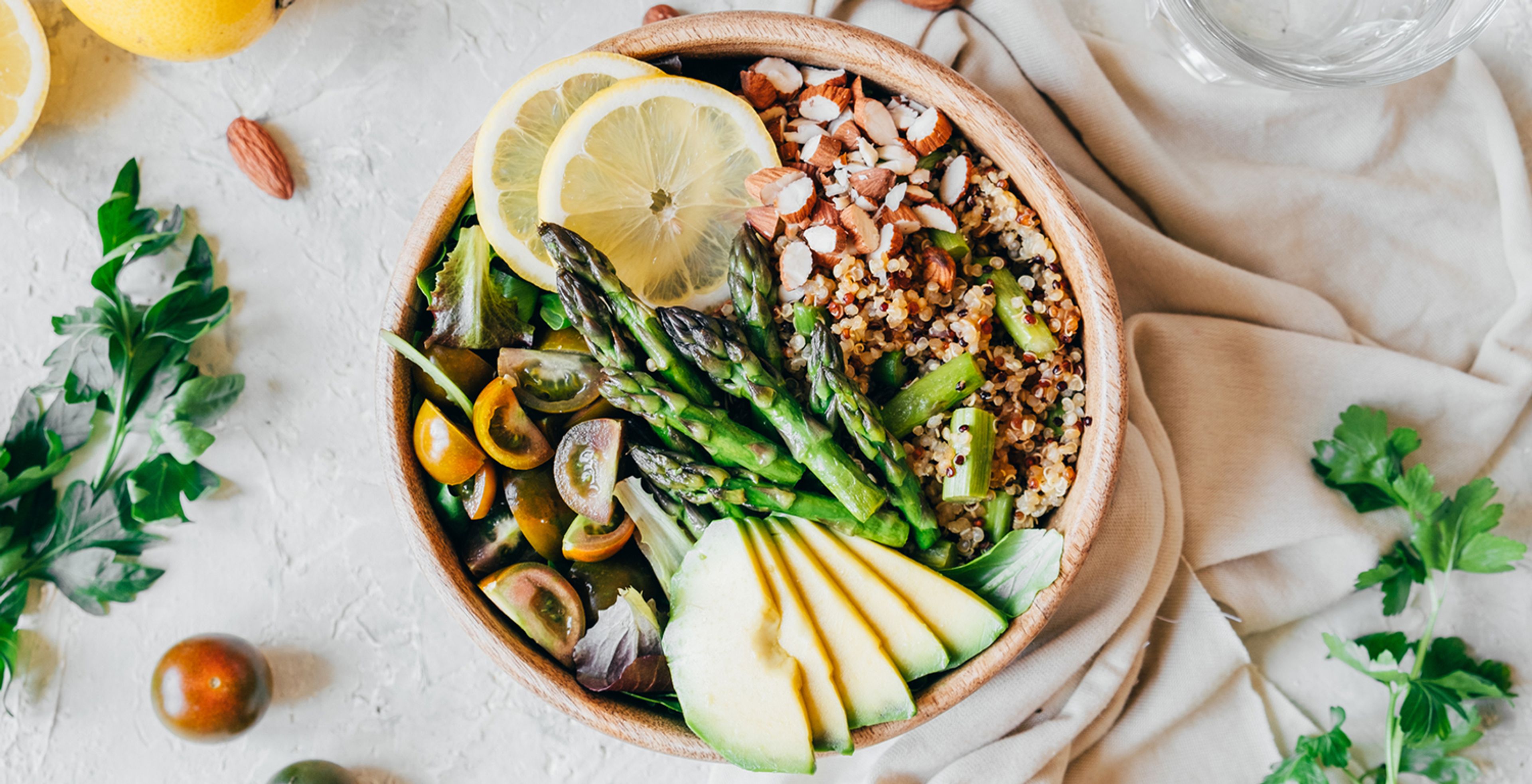 A wooden bowl filled with quinoa, asparagus, avocado slices, tomatoes, almond slivers and two slices of lemon on a bed of lettuce. On the sides of the bowl shows a cropped image of lemon slices, green garnishes, one almond, one tomato and a cropped glass of water.  