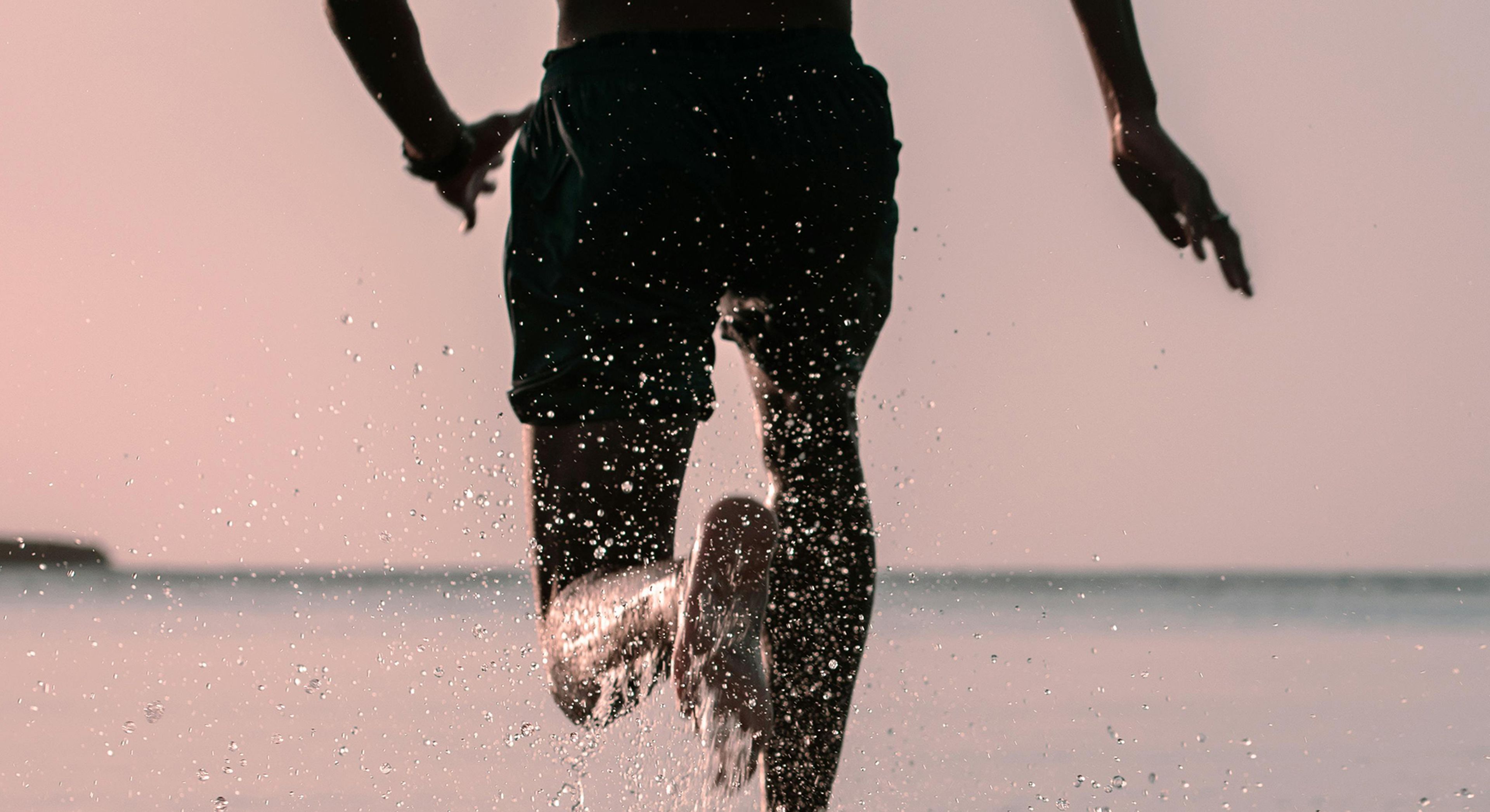 Man running with large body of water in the background, his left foot is splashing water