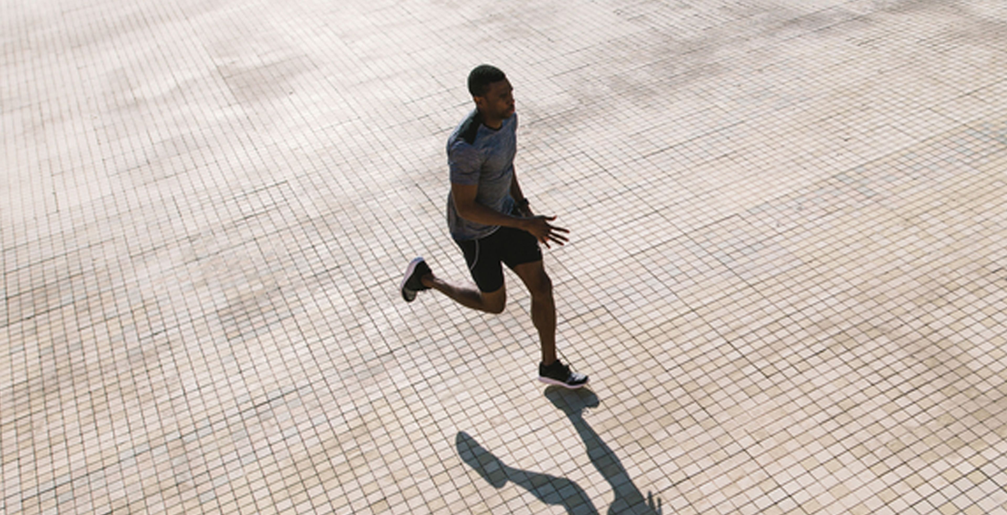 A Black man in a running pose, wearing black running shorts, dark colored sneakers and a gray exercise shirt.