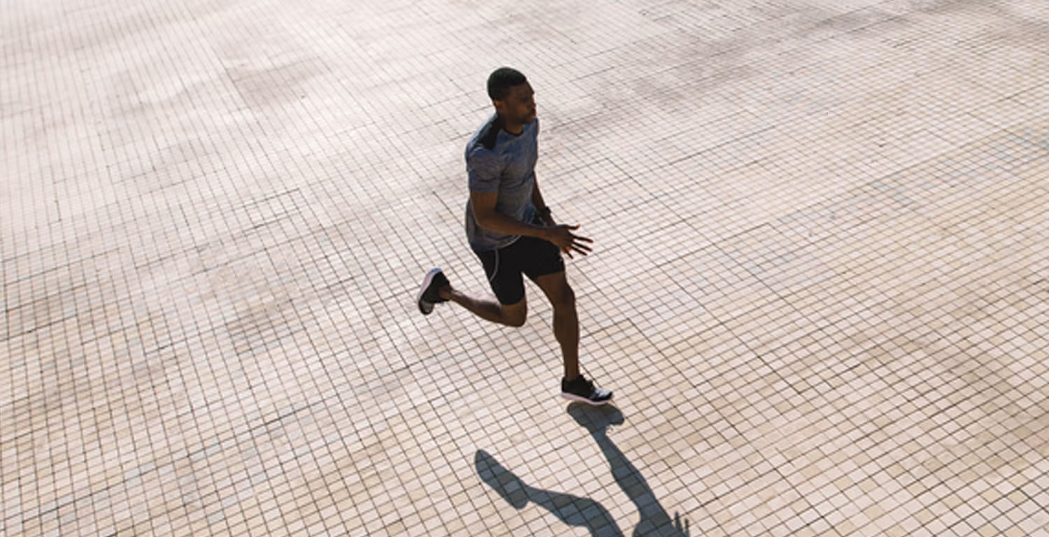 A Black man in a running pose, wearing black running shorts, dark colored sneakers and a gray exercise shirt.