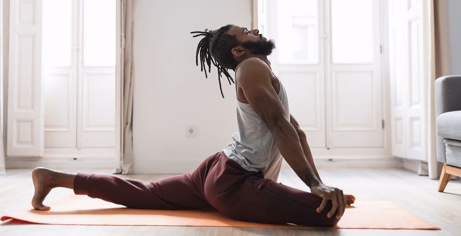 A Black man with dreads and a beard in pigeon pose, arches his back and looks up at the ceiling with his eyes closed. He is on an orange yoga mat, wearing red exercise pants and a gray exercise sleeveless top.