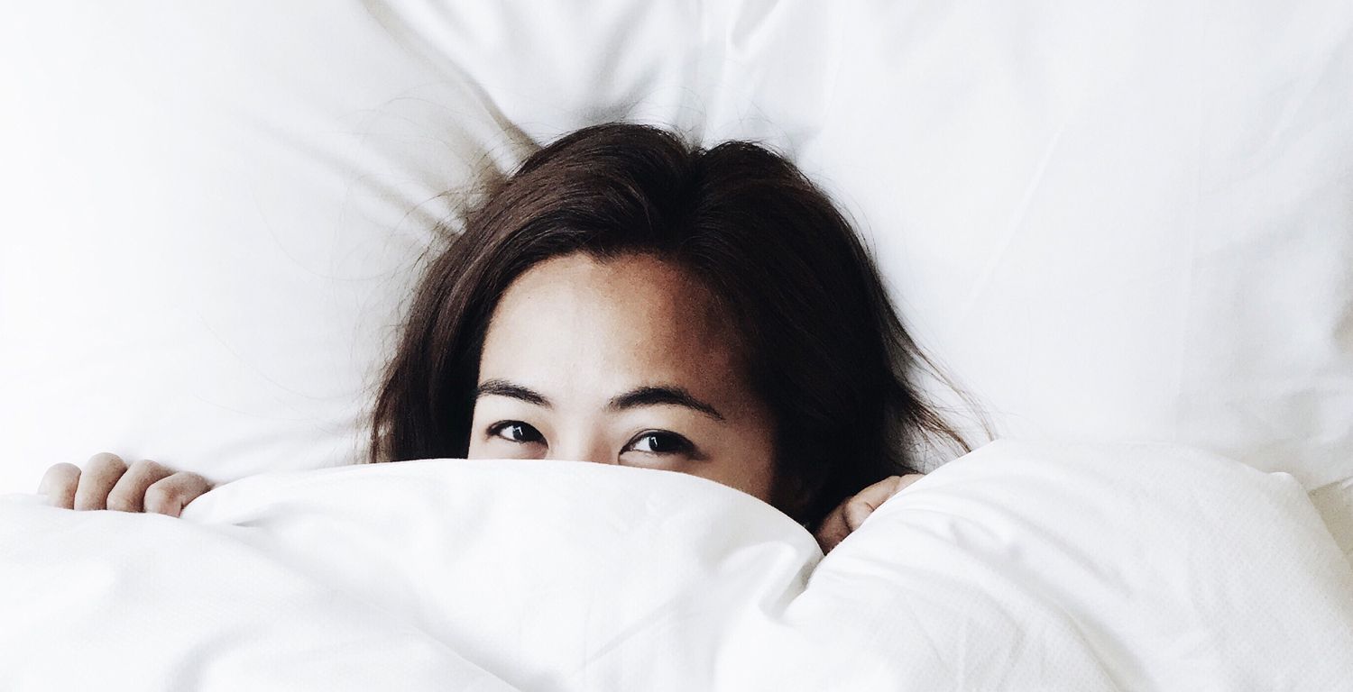 An Asian woman lying in a bed on a white pillow, covering her face with the white comforter revealing only her eyes, forehead and top of hair.