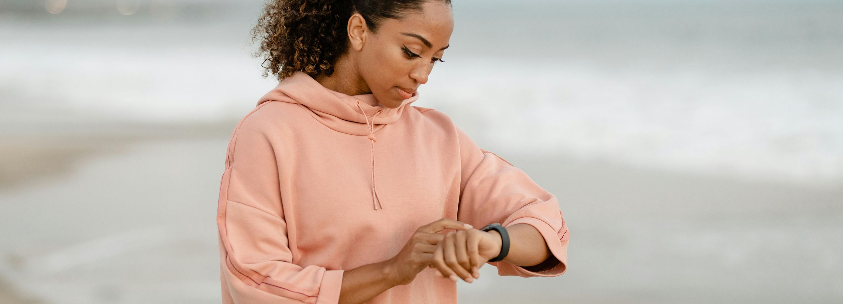 Woman on beach in a peach sweatshirt checking her smart watch