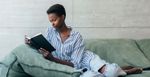 A black woman reclines on a sage green couch and reads a book. She has a short natural afro, wearing a white and blue striped blouse, ripped light denim jeans, white nail polish and a ring.