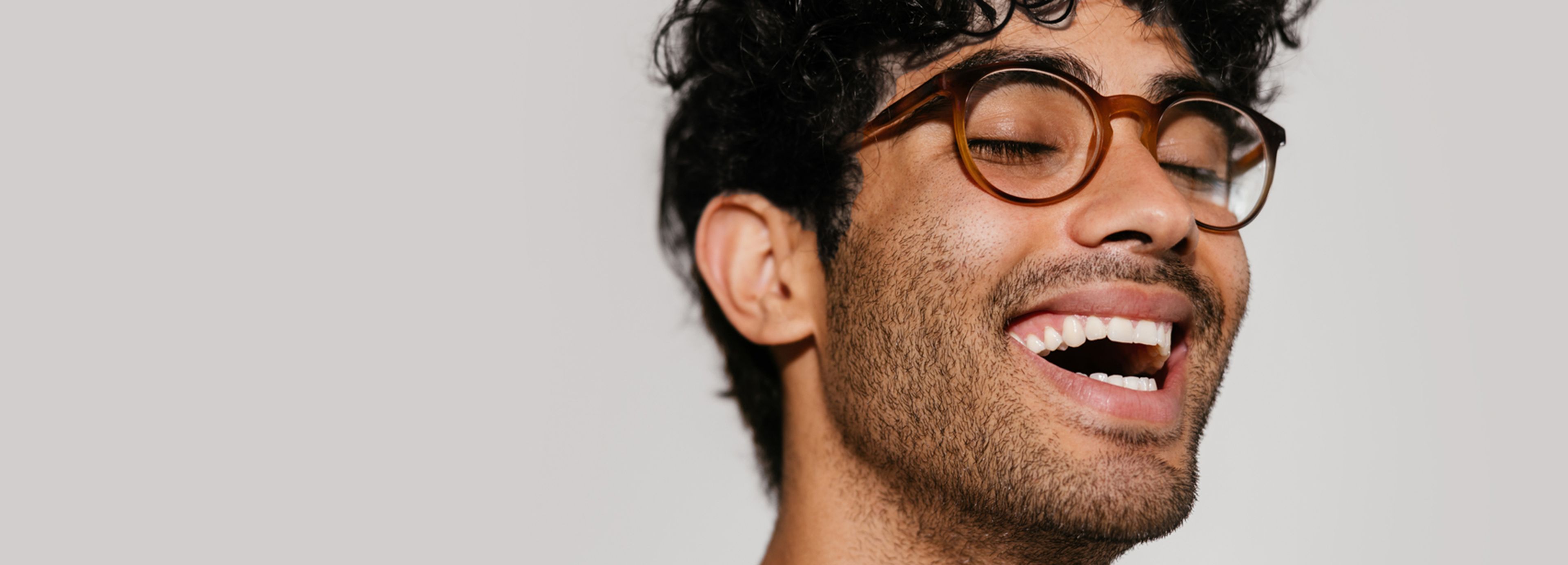 Young man with curly hair and round glasses laughing with eyes closed, expressing optimism joy, and emotional well-being.