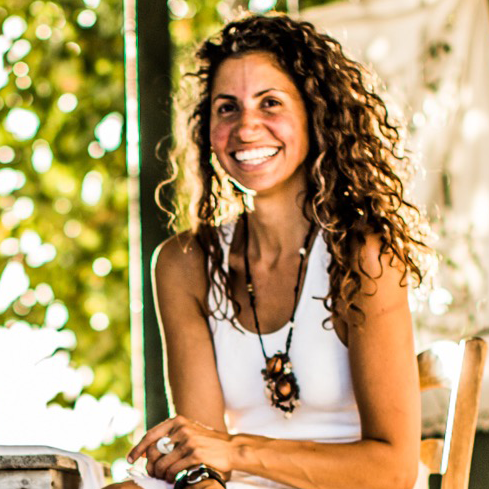 A tanned woman with brown curly hair. She is wearing a white tank top, long necklace, rings, smiling with teeth and looking forward.