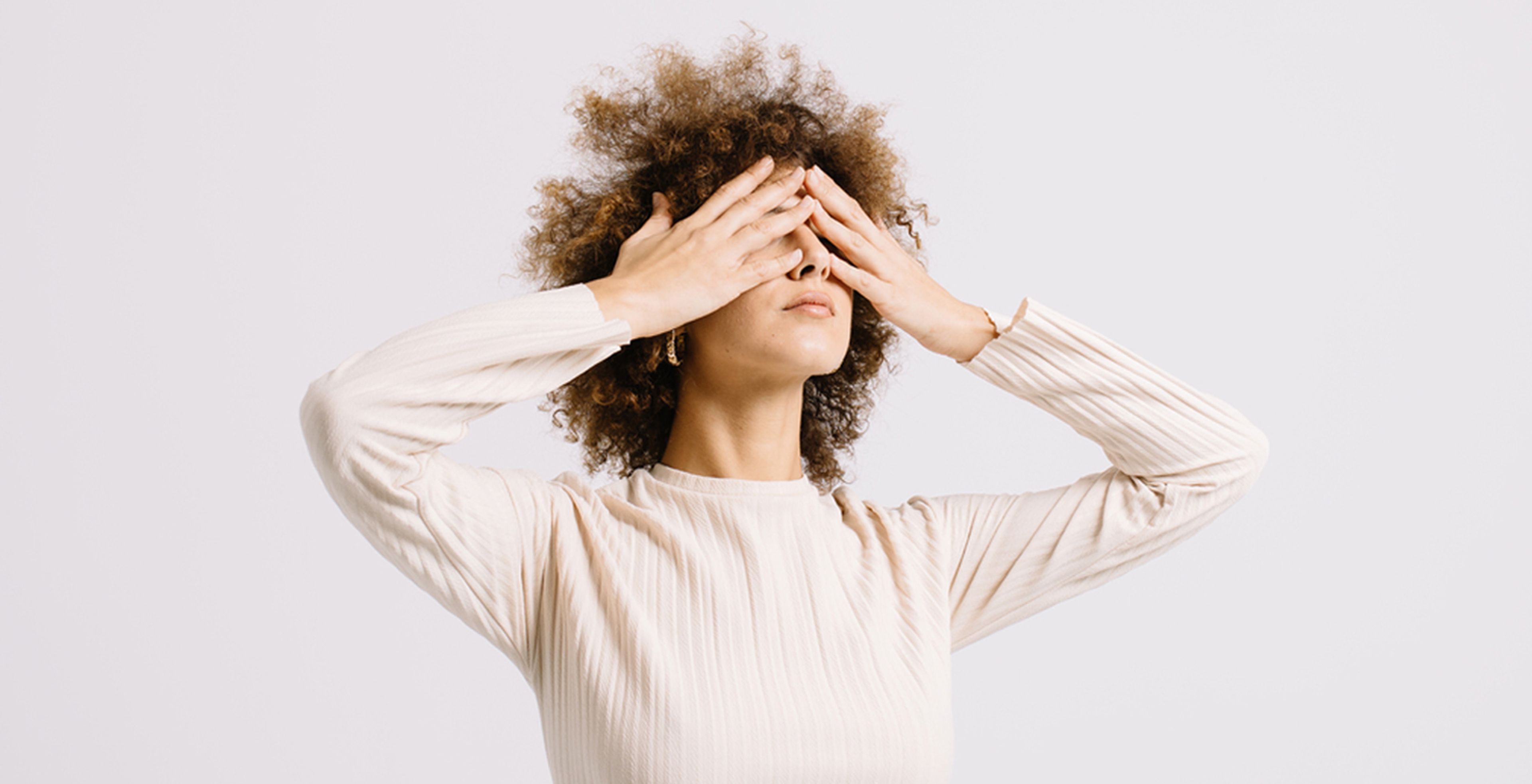 A light-skinned woman raises her arms and covers her eyes with both her hands. The woman has curly light brown hair, gold hoops and a cream-colored long sleeve shirt.