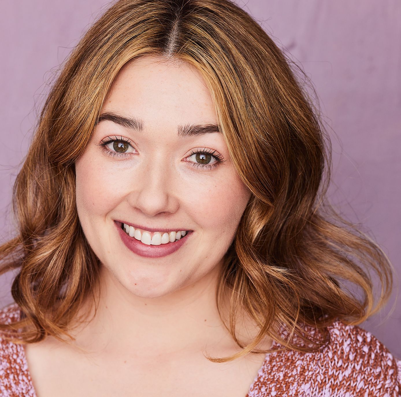 A white woman with blonde-brown hair and green eyes. She is wearing a v neck pink sweater and smiling with her front teeth showing.
