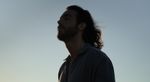 Man with long hair and beard looking up, covered by a shadow, against a blue sky