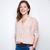A white woman in a pink blouse, brown belt, dark blue denim and a gold necklace. They have brown hair, blue eyes and pink lipstick. They have one arm behind their back and another hand in their jean pocket, smiling with teeth looking forward. 