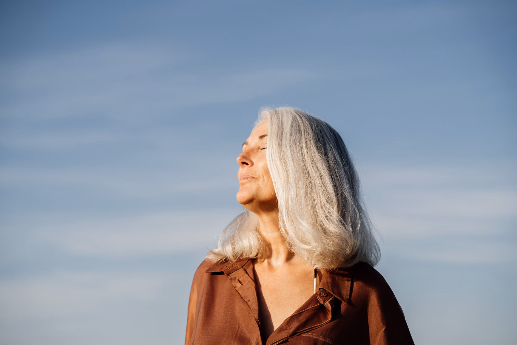 Grey haired woman with eyes closed, standing in front of a blue sky