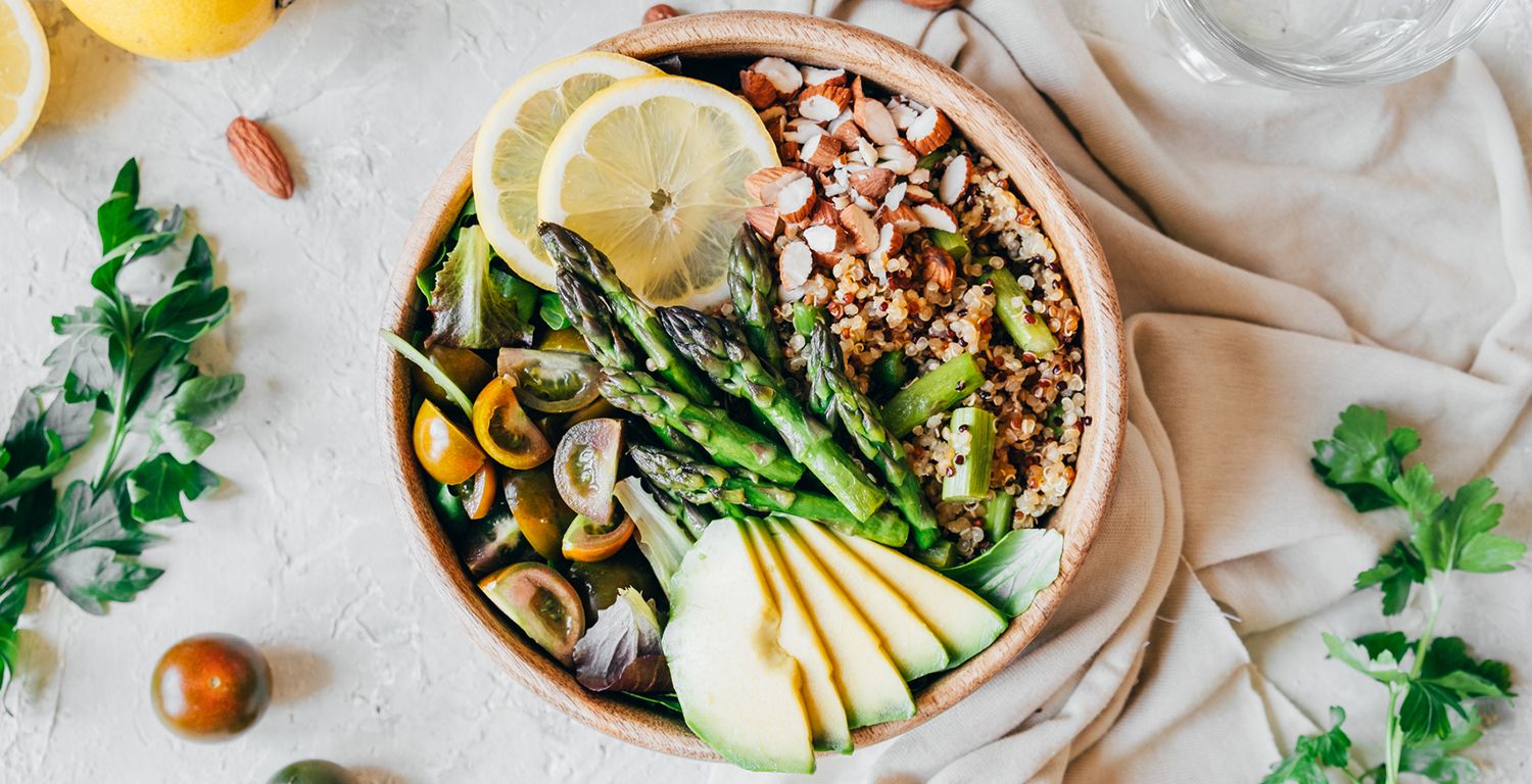 A wooden bowl filled with quinoa, asparagus, avocado slices, tomatoes, almond slivers and two slices of lemon on a bed of lettuce. On the sides of the bowl shows a cropped image of lemon slices, green garnishes, one almond, one tomato and a cropped glass of water.