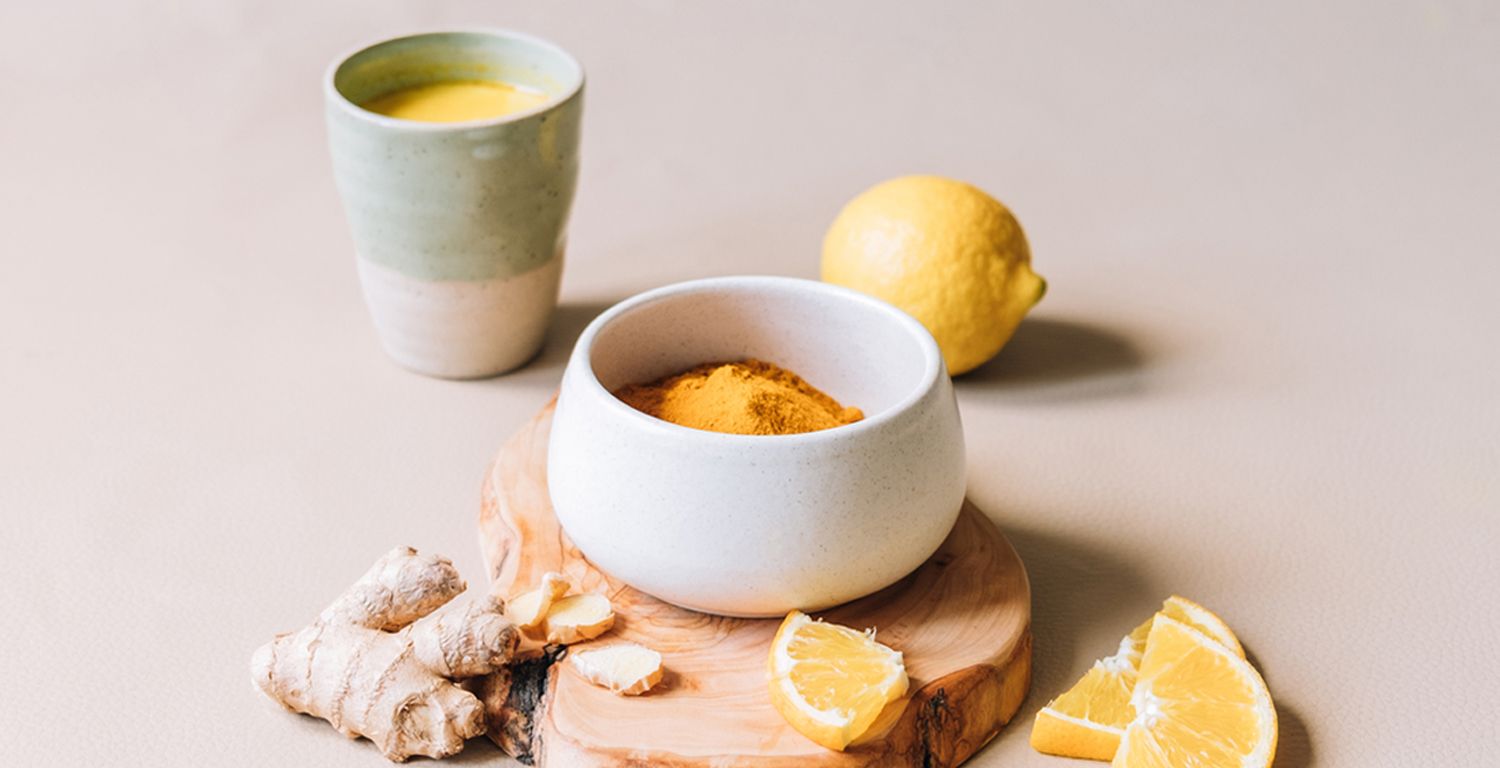 A small ceramic bowl with turmeric rests on a piece of wood, surrounded by ginger root, lemon slices and a ceramic cup full of yellow/orange liquid.