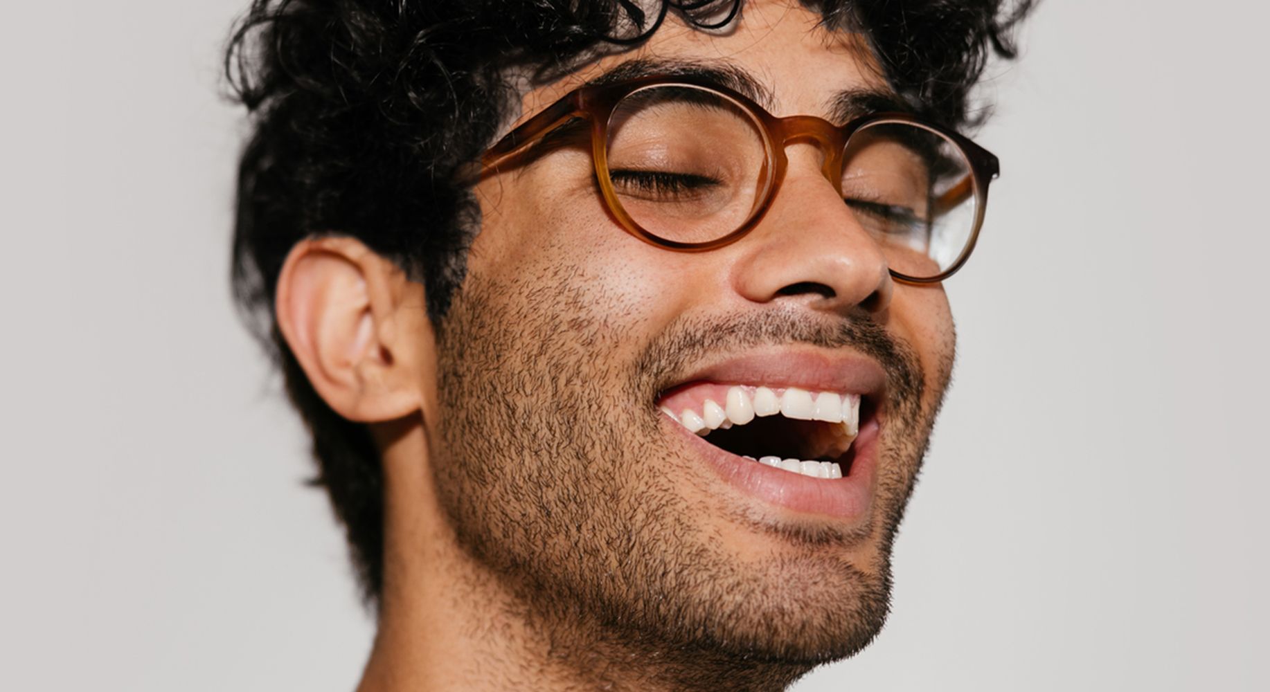 Young man with curly hair and round glasses laughing with eyes closed, expressing optimism joy, and emotional well-being.