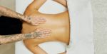 A massage therapist places their white hands on the back of a light-skinned naked woman, laying down on a massage table. A blanket is covering her bottom half and you can see the massage therapist's tattooed arms too.