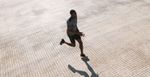 A Black man in a running pose, wearing black running shorts, dark colored sneakers and a gray exercise shirt.