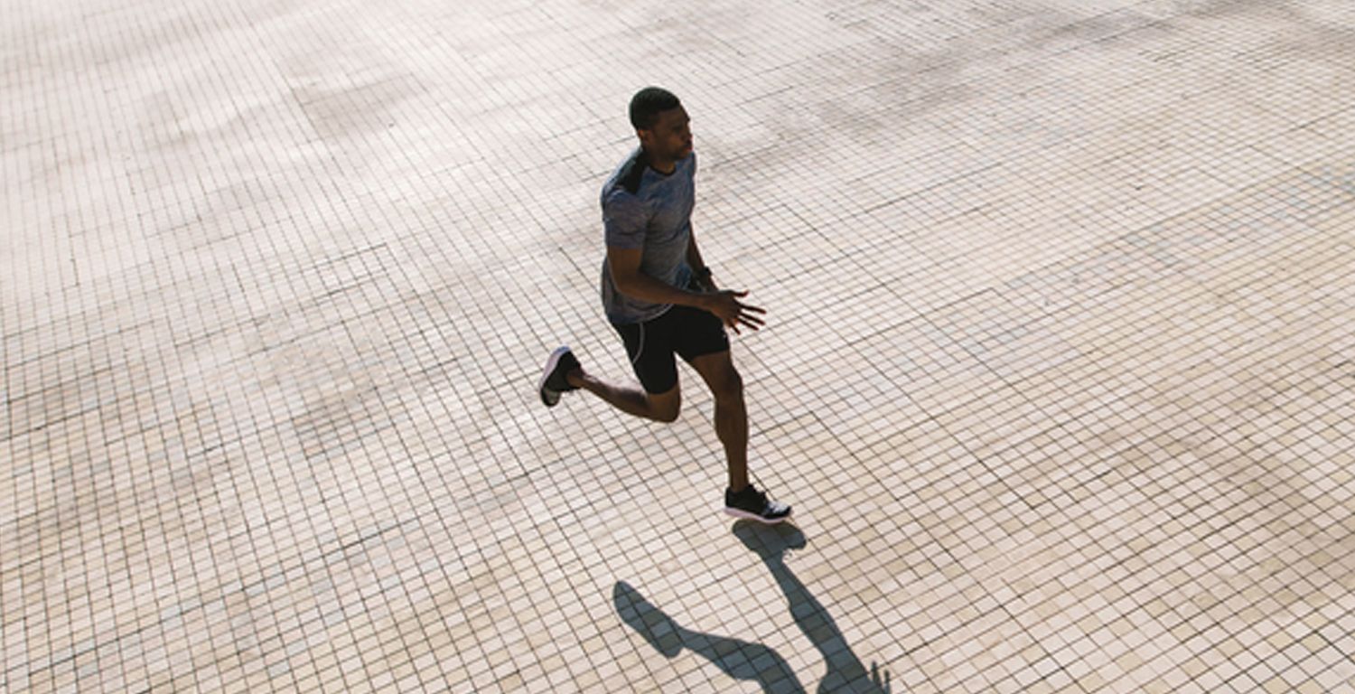 A Black man in a running pose, wearing black running shorts, dark colored sneakers and a gray exercise shirt.