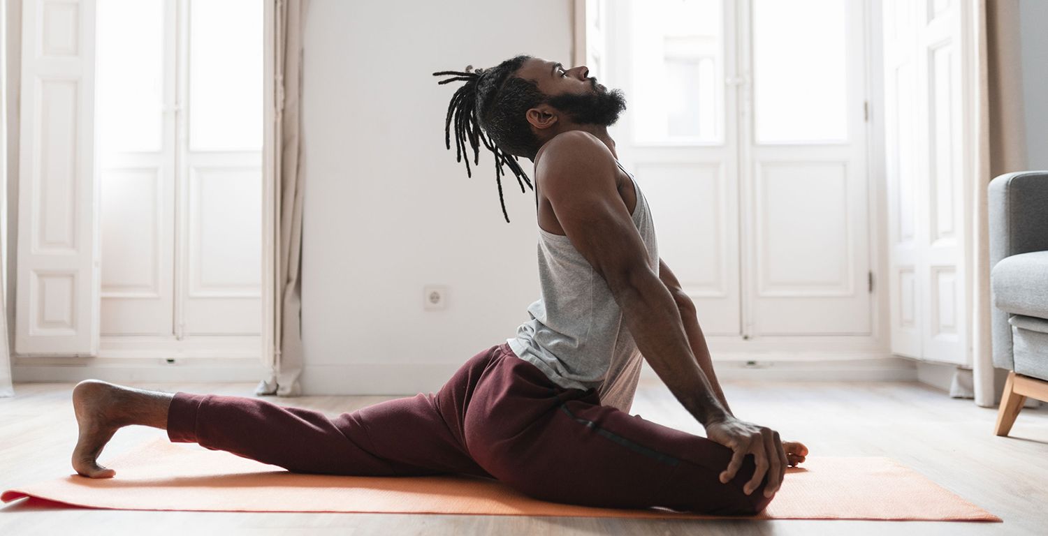 A Black man with dreads and a beard in pigeon pose, arches his back and looks up at the ceiling with his eyes closed. He is on an orange yoga mat, wearing red exercise pants and a gray exercise sleeveless top.