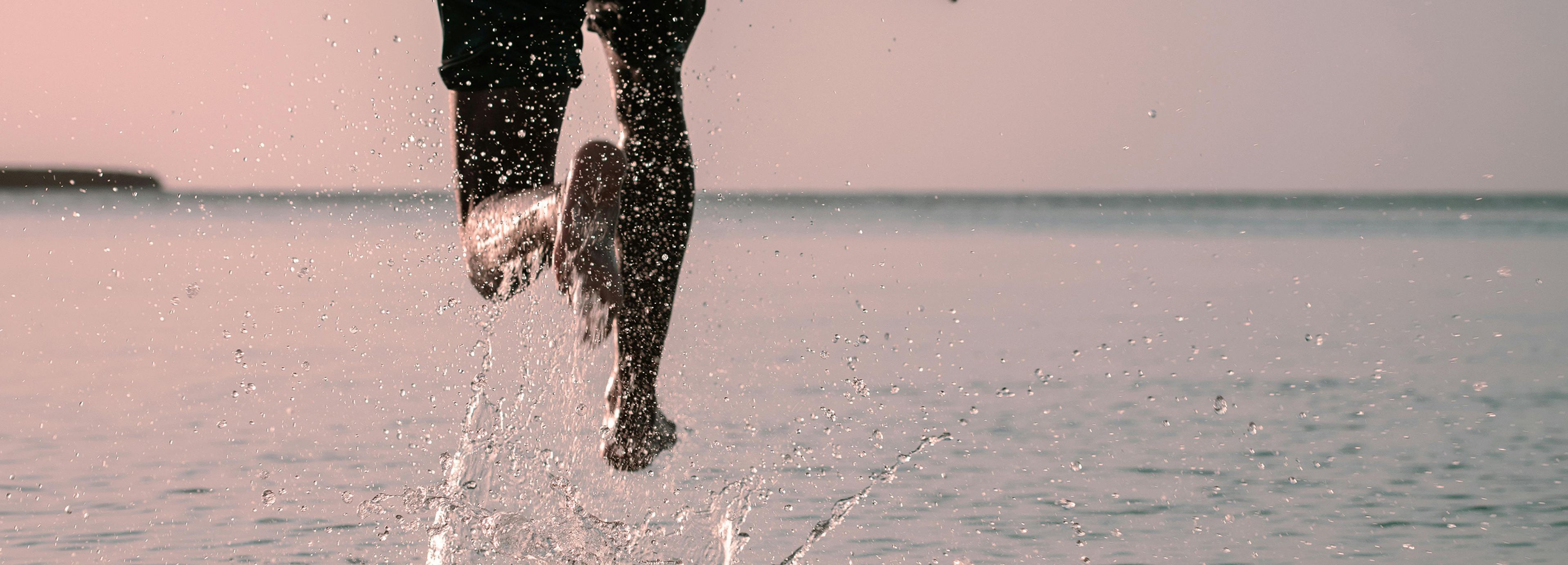 Man running with large body of water in the background, his left foot is splashing water