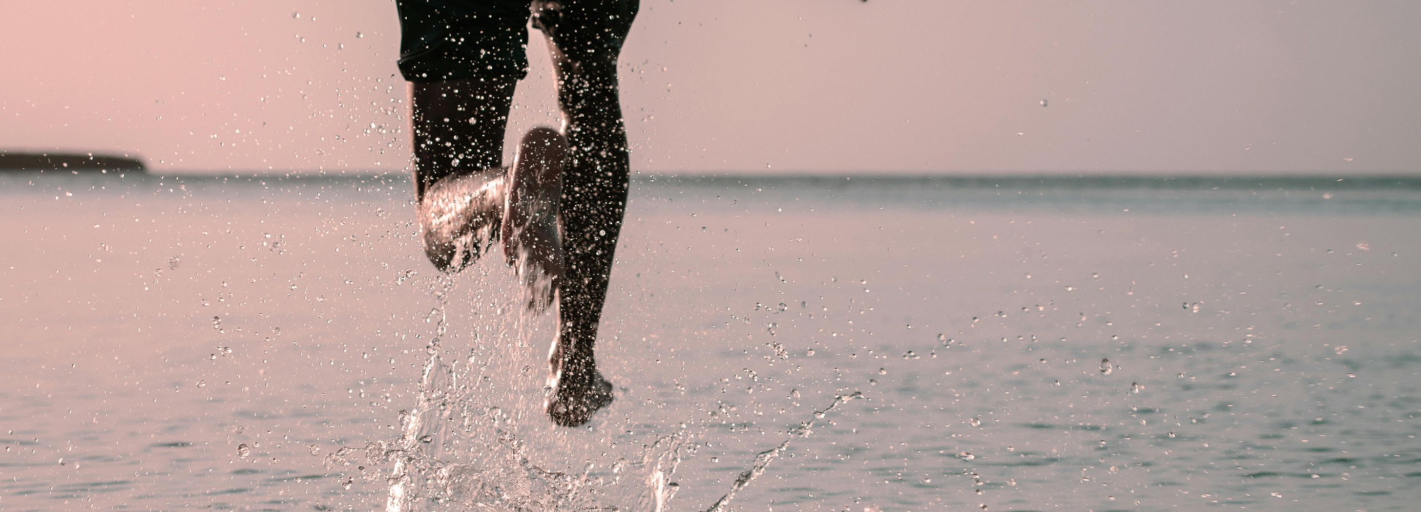 Man running with large body of water in the background, his left foot is splashing water