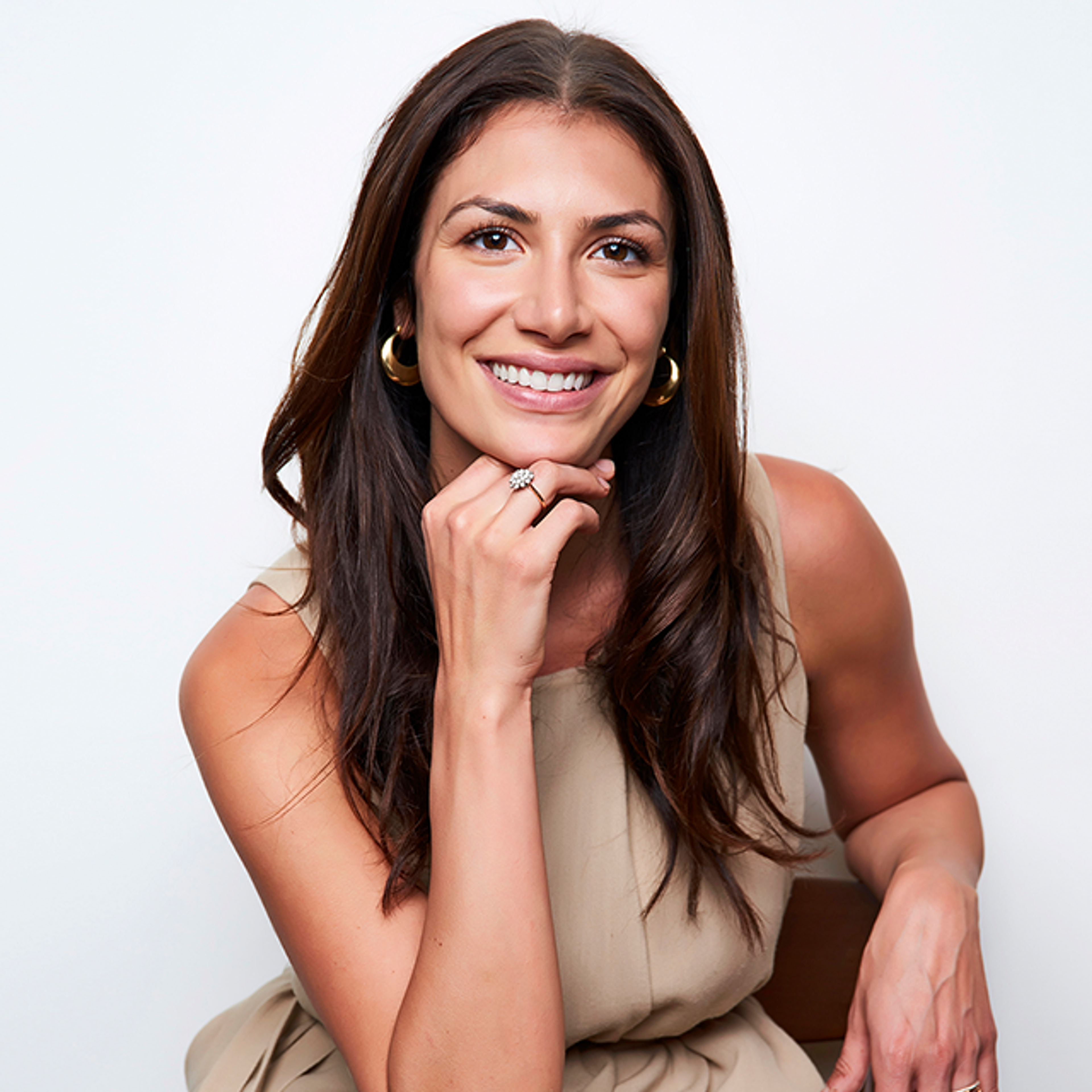 An image of Lia Avellino, LCSW. She has medium-length dark brown hair, brown eyes and olive skin. She is wearing a tan dress, gold earrings and a wedding ring. She is leaning over with her hand rested on her chin, while her other arm is resting on the chair she is sitting on. She is smiling and looking forward.