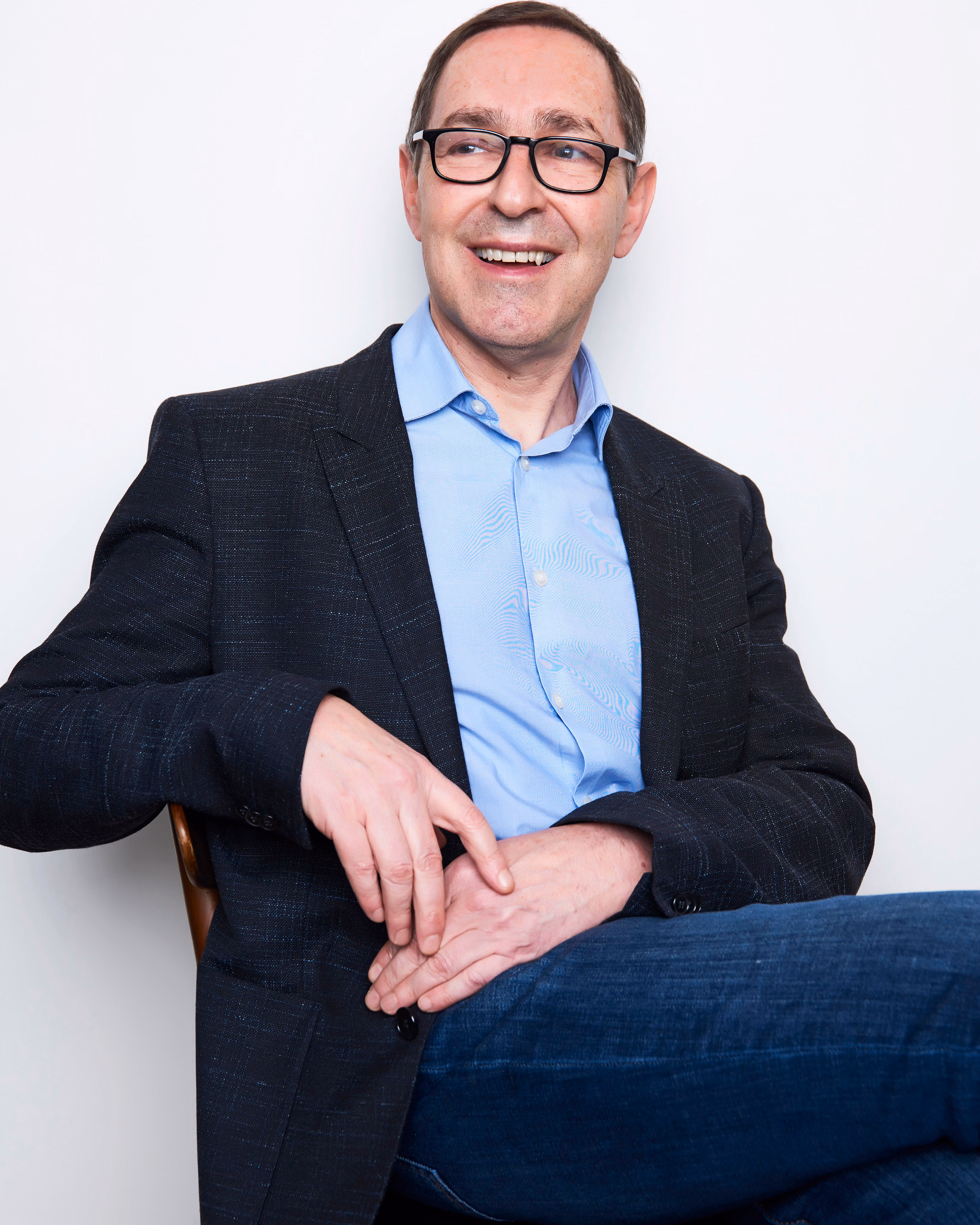 Dr. Frank Lipman sits on a chair, cross-legged, arm resting on the back of the chair, lightly touching his other hand that is resting on his thigh. He is wearing blue denim, a blue dress shirt and a navy textured blazer and black glasses. He is smiling, showing his front teeth looking off to the side.