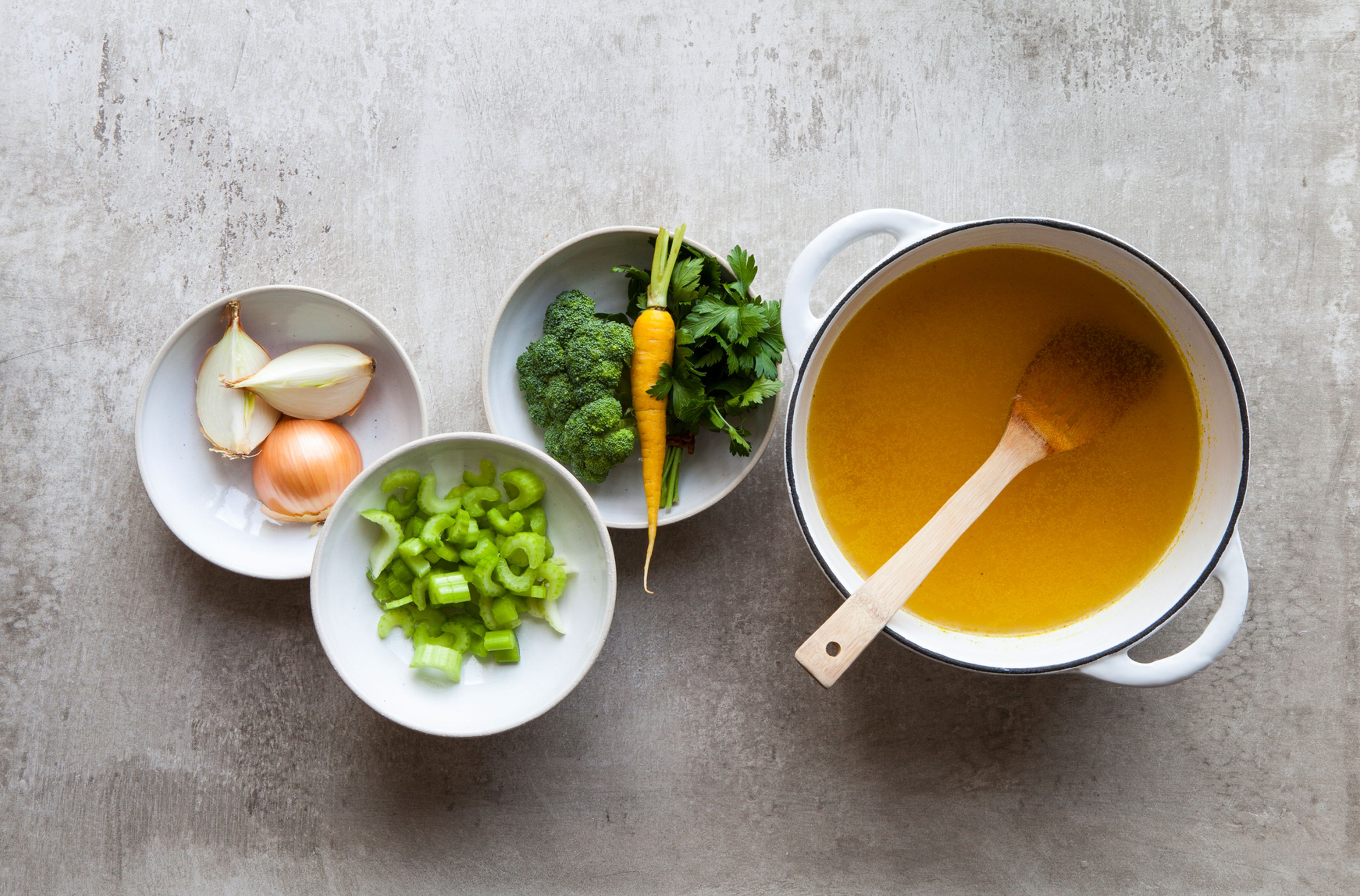 pot of bone broth with bowls of celery, broccoli, carrots, onions