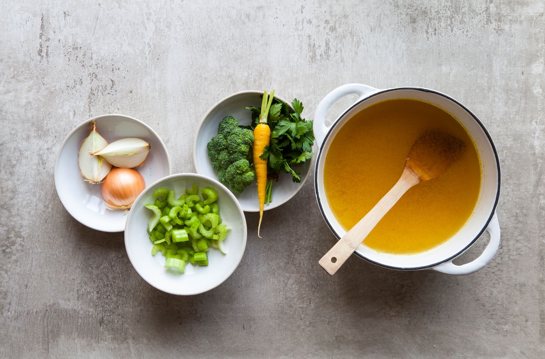 pot of bone broth with bowls of celery, broccoli, carrots, onions