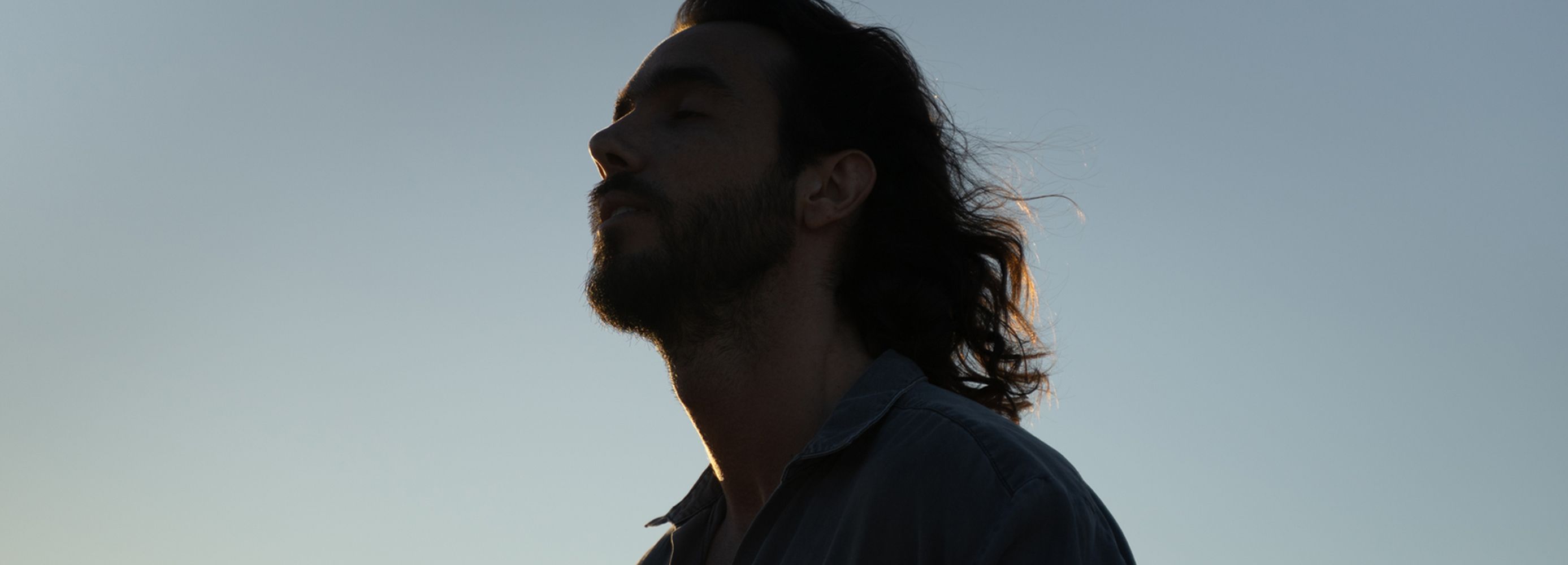 Man with long hair and beard looking up, covered by a shadow, against a blue sky