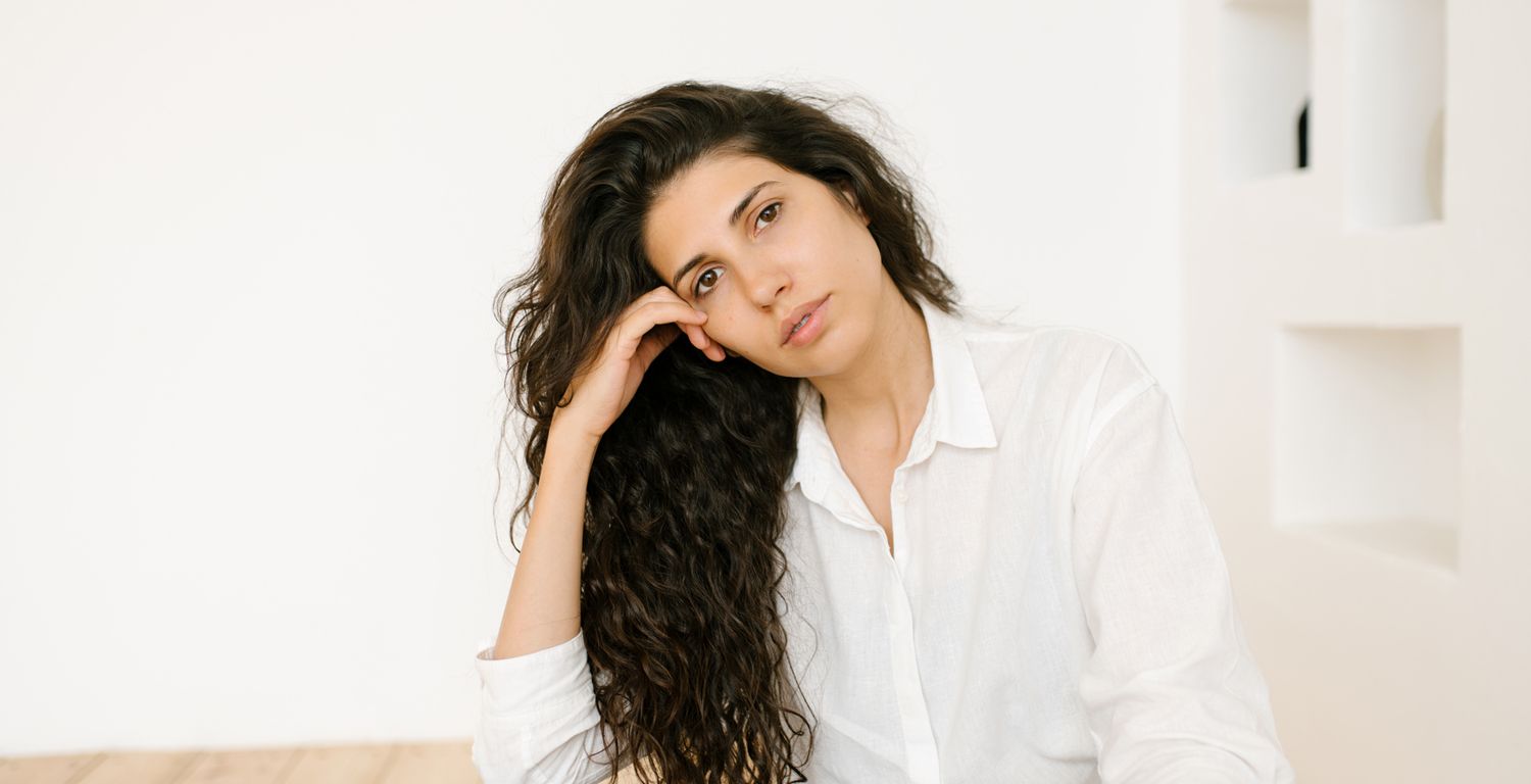 A White woman with long curly brown hair, rests her head on her hand, looking in the distance.