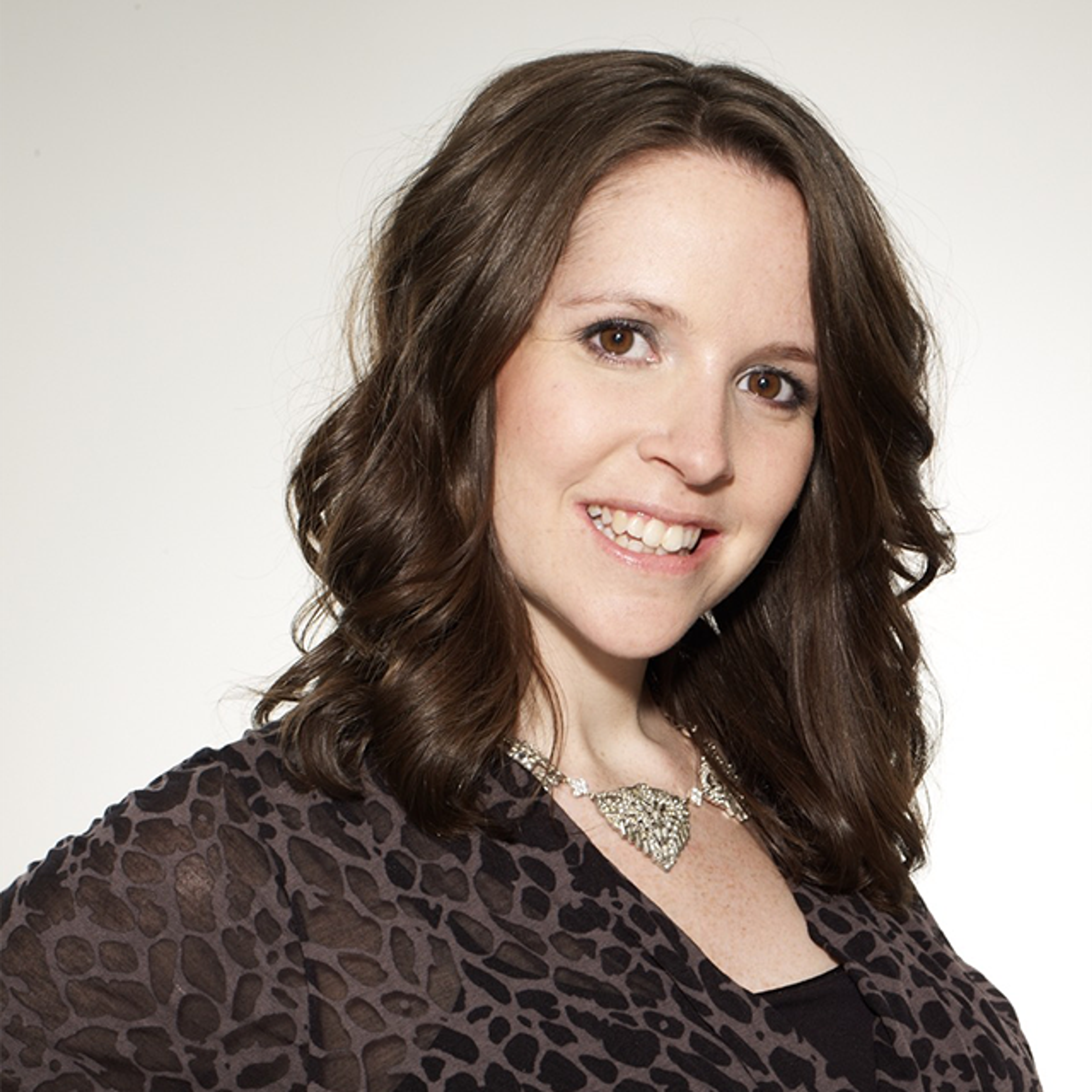 A white woman with dark brown curly hair and brown eyes. She is wearing a black, animal printed blouse, silver necklace and smiling with teeth, looking forward.