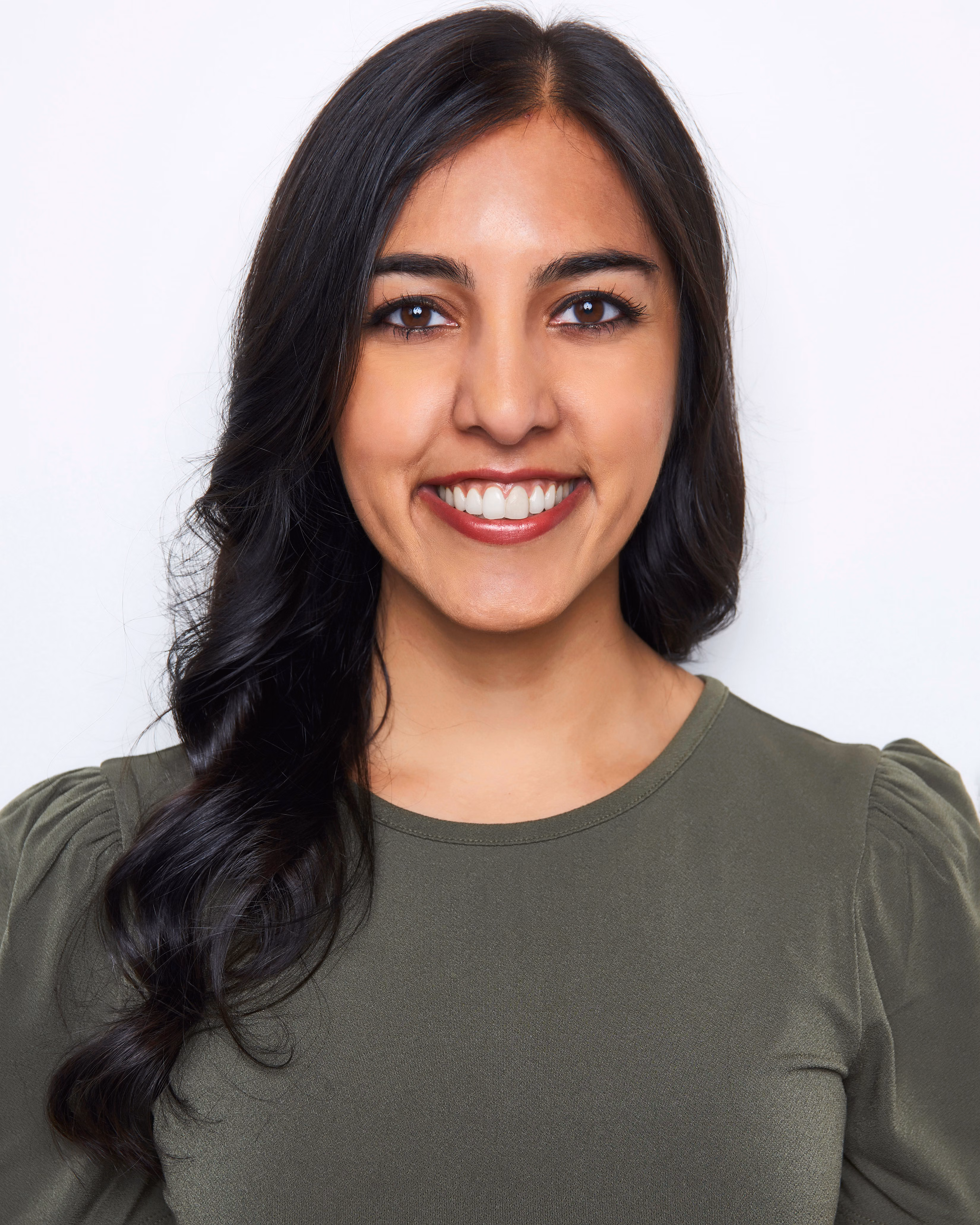 An Indian woman with red lipstick, brown eyes, dark brown hair and wearing a green shirt. She is smiling with her front teeth showing, looking forward.