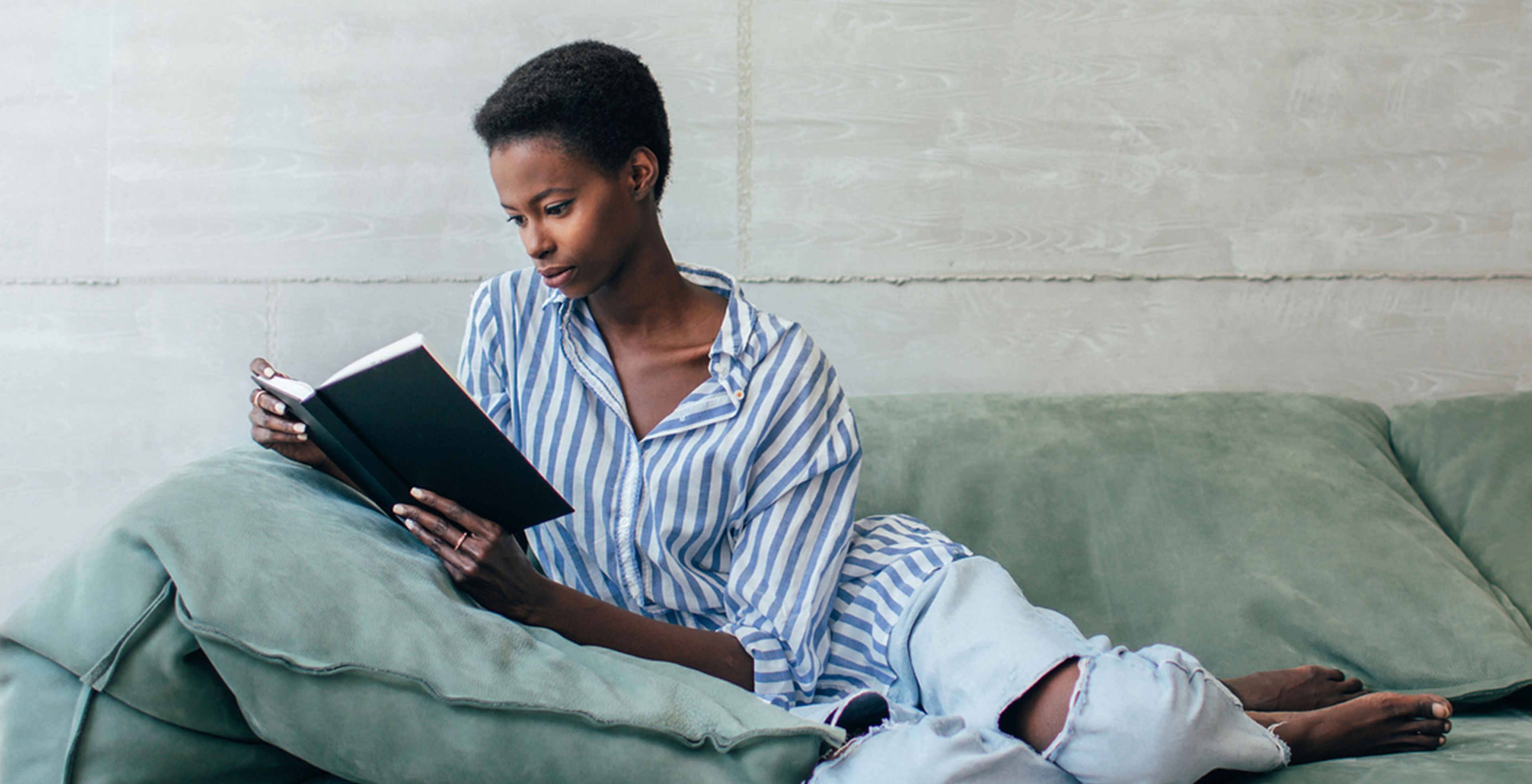 A black woman reclines on a sage green couch and reads a book. She has a short natural afro, wearing a white and blue striped blouse, ripped light denim jeans, white nail polish and a ring.